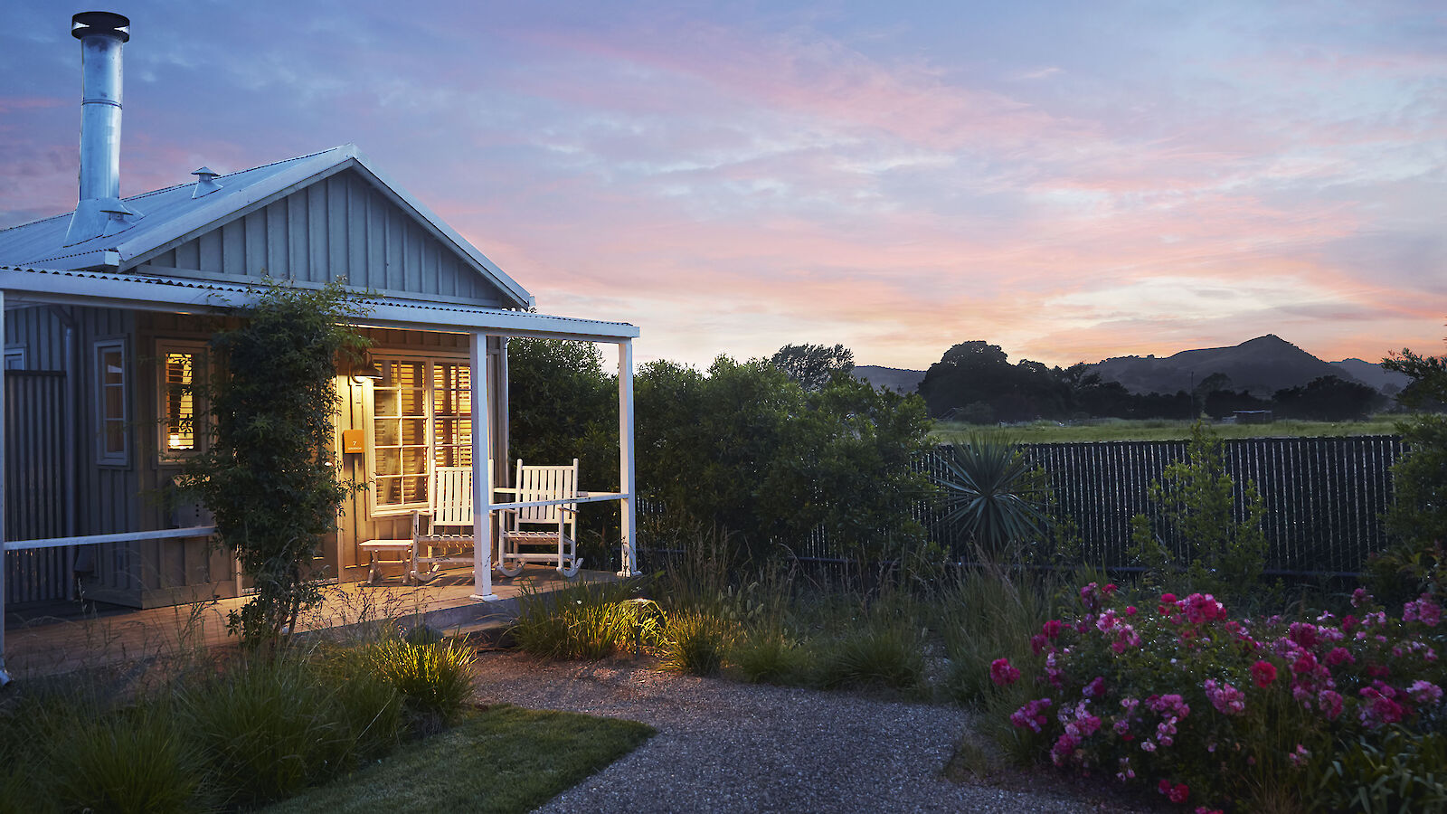 A cozy cottage with a porch is surrounded by greenery and flowers under a colorful sunset sky.