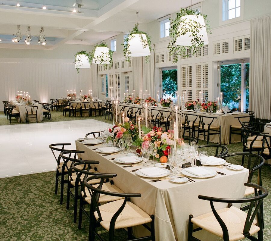 An elegantly decorated dining hall with arranged tables, floral centerpieces, and hanging lights with greenery, ready for an event.