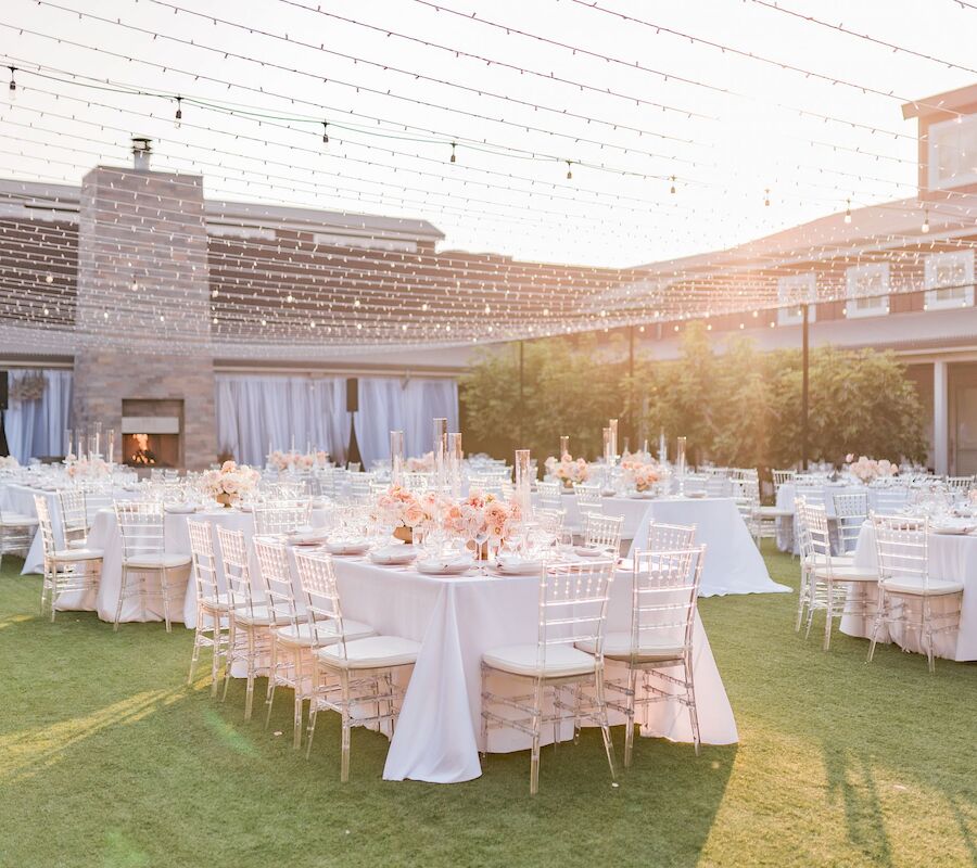 An elegant outdoor event setup features round tables with white tablecloths and decor, surrounded by white chairs, under string lights.