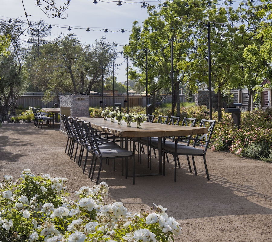 A long outdoor table surrounded by empty chairs is set in a garden with string lights overhead and blooming flowers along the pathway.