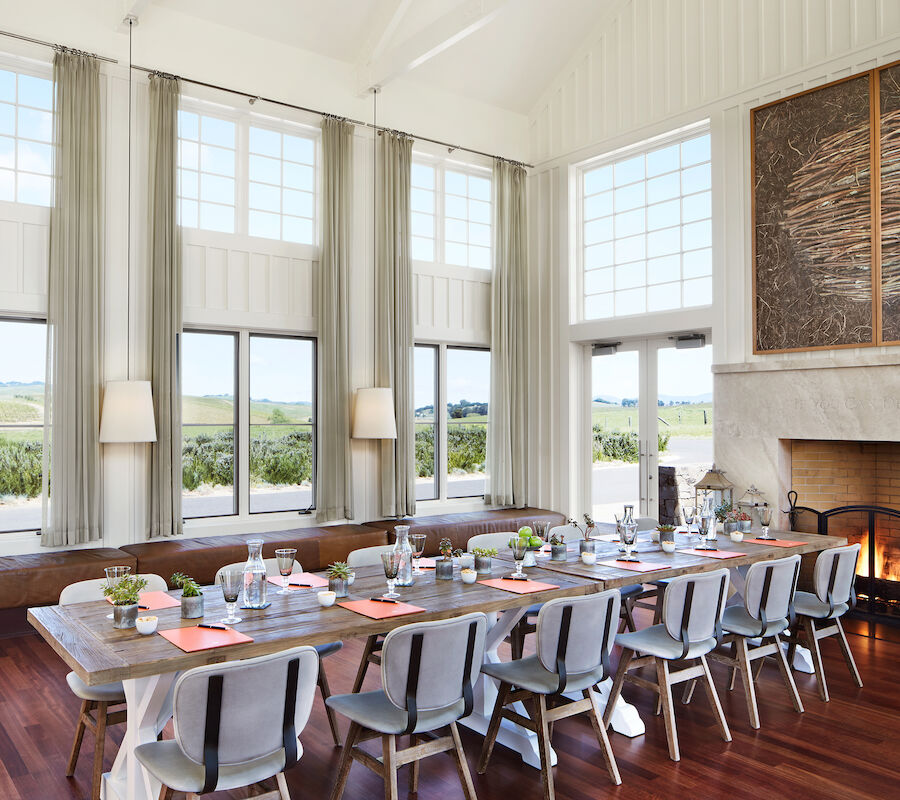 A spacious dining room with a long table, modern chairs, large windows, a fireplace, and a scenic outdoor view. The setting appears elegant and inviting.