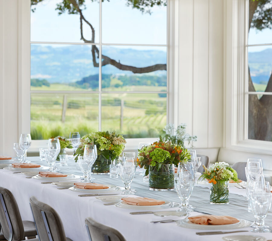 A dining setup with a long table is elegantly decorated with flowers and table settings in front of large windows showcasing a scenic countryside view.